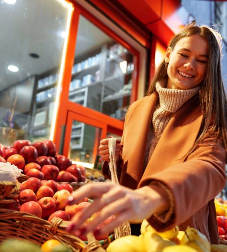 Good looking young woman standing in front of fruit shelves buying groceries in the street close up